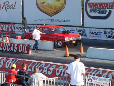 2003 Ford Crown Victoria - man directs a red gasser-style classic car staging at a drag strip beside orange cones and sponsor banners.