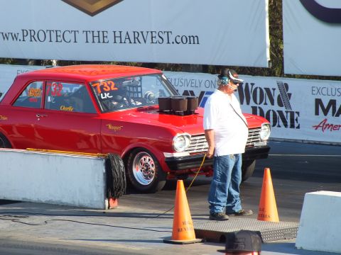 2003 Ford Crown Victoria - man stands beside a red two-door drag car marked 732 at the staging line, holding a cord among orange cones.