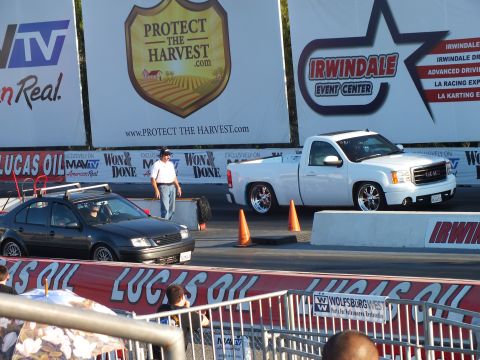 2003 Ford Crown Victoria - lowered white pickup and a black sedan stage at a drag strip beneath Irwindale, MAVTV and Protect the Harvest banners.