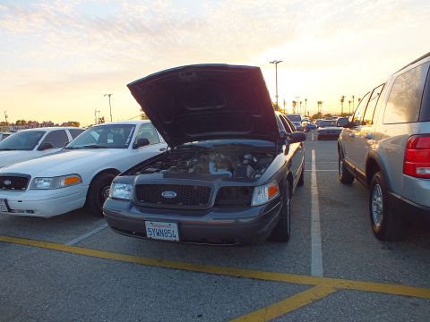 2003 Ford Crown Victoria - black Ford sedan with its hood up sits between a white sedan and a silver SUV in a parking lot at sunset.