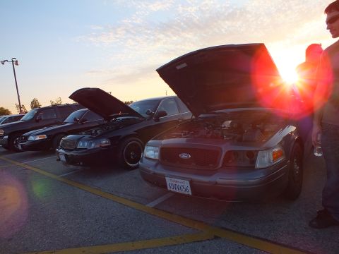 2003 Ford Crown Victoria - Two dark Ford sedans with raised hoods are backlit by the setting sun in a parking lot, a man standing at right.