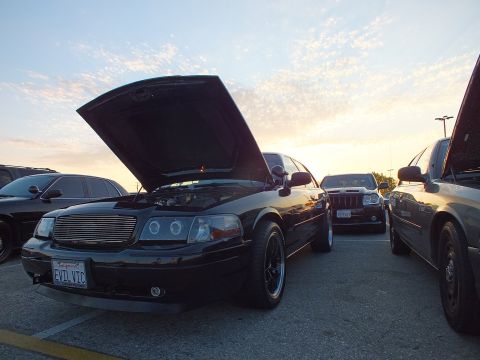2003 Ford Crown Victoria - black sedan with a billet grille and raised hood, plate reading EVIL VIC, sits in a parking lot at sunset.