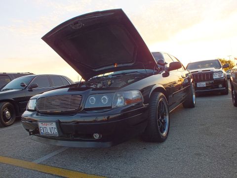 2003 Ford Crown Victoria - lowered black sedan with a billet grille, black wheels and hood up, plate EVIL VIC, parked in a lot at sunset.