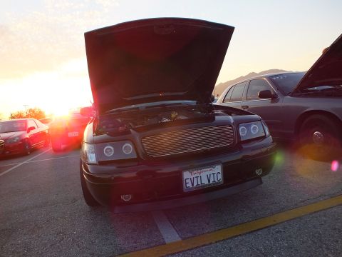 2003 Ford Crown Victoria - Front view of a black sedan with a billet grille and raised hood, plate reading EVIL VIC, backlit at sunset.