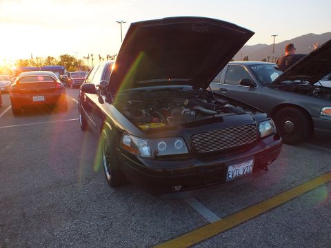 2003 Ford Crown Victoria - black sedan with raised hood and billet grille, plate EVIL VIC, sits in a sun-flared parking lot among other cars.
