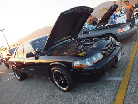 2003 Ford Crown Victoria - modified black full-size sedan sits with its hood open at a sunset car-show meet, mountains in the background.