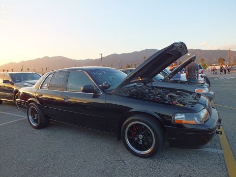 2003 Ford Crown Victoria - black full-size sedan with raised hood and black wheels parked at a car meet with mountains behind.