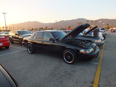 2003 Ford Crown Victoria - black full-size sedan with its hood open sits among parked cars at a sunset car-show lot.
