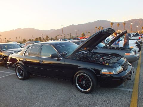 2003 Ford Crown Victoria - Front three-quarter view of a black full-size sedan with open hood, palm trees and mountains behind.