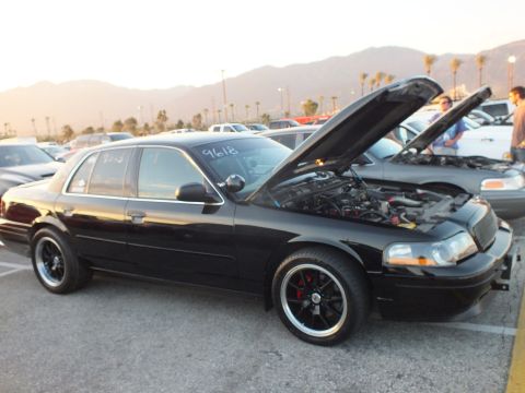 2003 Ford Crown Victoria - black full-size sedan with raised hood and black multi-spoke wheels at a sunset car meet.