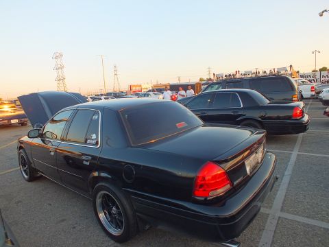 2003 Ford Crown Victoria - black full-size sedan viewed from the rear among parked cars and SUVs at a sunset car show.