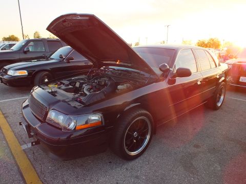 2003 Ford Crown Victoria - black full-size sedan with its hood open, backlit by the setting sun at a car meet.