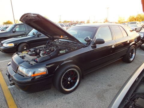 2003 Ford Crown Victoria - black full-size sedan with open hood revealing the engine bay, beside another sedan at sunset.