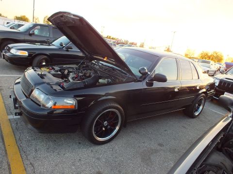 2003 Ford Crown Victoria - black full-size sedan with raised hood and black wheels, backlit by sunset at a car meet.