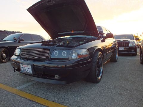 2003 Ford Crown Victoria - black Ford Crown Victoria with halo headlights, open hood, and a California 'EVIL VIC' plate at sunset.