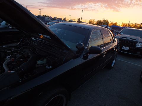 2003 Ford Crown Victoria - black sedan with its hood up is shown side-on at sunset, parked beside a dark SUV in a lot under a colorful sky.