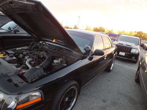 2003 Ford Crown Victoria - black sedan with raised hood reveals its engine and supercharger area, riding on black wheels in a parking lot at dusk.