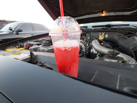 2003 Ford Crown Victoria - red slushie in a clear domed cup sits on a car's cowl panel, with the open engine bay and another vehicle visible behind it.