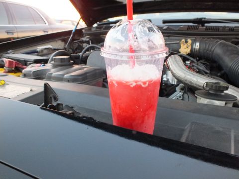 2003 Ford Crown Victoria - red icy beverage in a domed plastic cup rests on a black car's cowl, with the engine compartment and a sedan in the background.