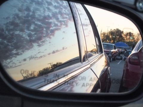 2003 Ford Crown Victoria - side-mirror reflection captures a chrome-trimmed sedan against a sunset sky, with parked cars and a blue canopy in the lot.