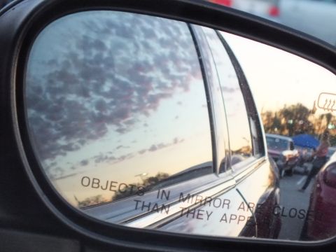 2003 Ford Crown Victoria - side mirror reflects a chrome-trimmed sedan and a cloud-filled sunset, with the etched 'Objects in mirror' warning visible.