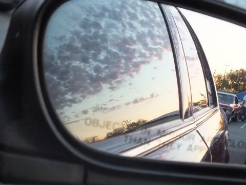 2003 Ford Crown Victoria - side mirror reflects a dark sedan and pink-streaked sky, with people and parked cars filling the lot in the background.