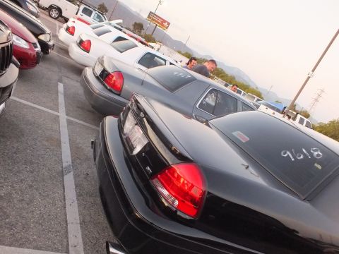 2003 Ford Crown Victoria - black sedan's rear is marked with '9618' in window chalk as it sits among parked cars in a lot at dusk with mountains behind.