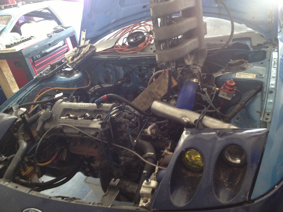 Engine bay of a blue car with aftermarket intake and turbo components. Another vehicle is partially visible in the background
