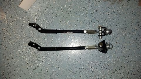 1970 Ford Torino - Two black adjustable suspension control arms are laid out on a speckled gray surface, showing their metal components and mounting points.