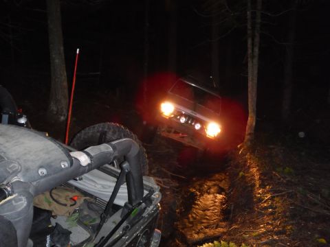 2005 Jeep Wrangler - Two off-road vehicles navigate a muddy trail through a forest at night, illuminated by headlights.