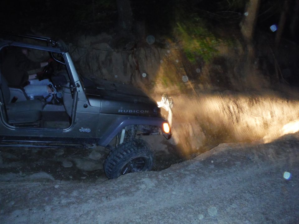 Jeep Wrangler driving on a dirt trail at night, headlights on, going through a narrow passage