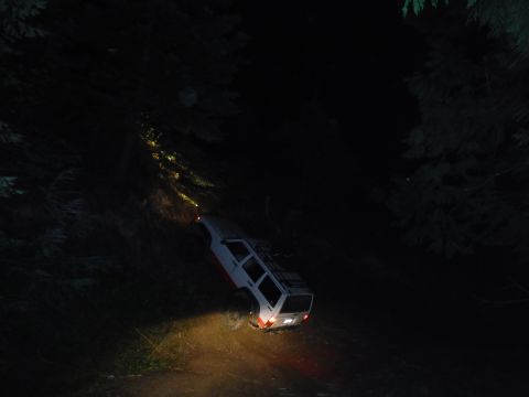 2005 Jeep Wrangler - A white Jeep Cherokee navigates a steep, dark, wooded trail at night, with its headlights illuminating the path.