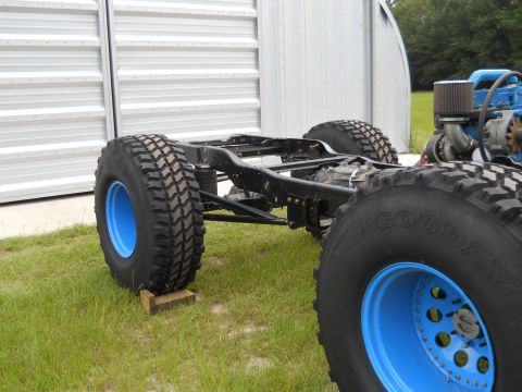 1992 Ford Bronco - The chassis and engine of a vehicle are shown outdoors, with large off-road tires mounted on blue wheels.