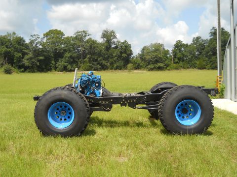 1992 Ford Bronco - A truck chassis with an engine and oversized tires sits on a grassy field under a cloudy sky.