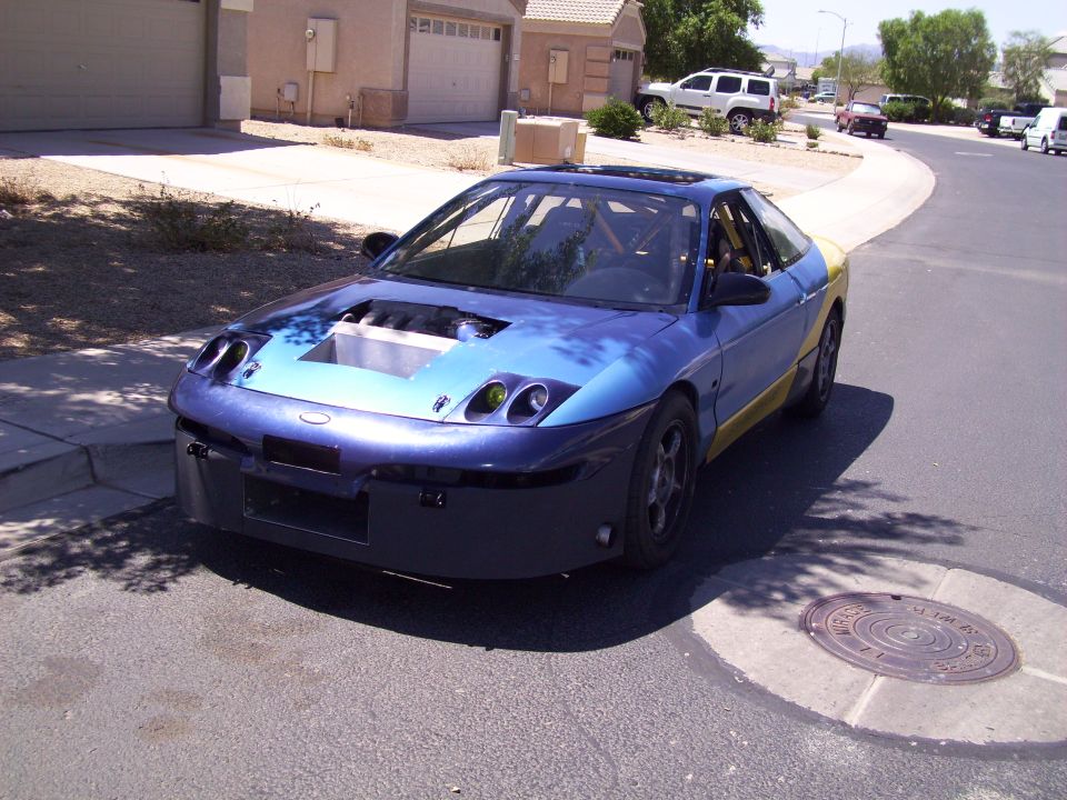 Blue and yellow coupe with hood removed, showing engine bay. Roll cage visible inside