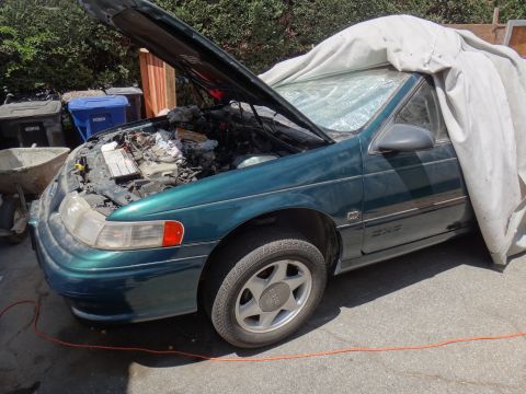 1995 Ford Taurus - A green sedan sits outside with its hood open, revealing the engine bay, and is partially covered by a tarp.