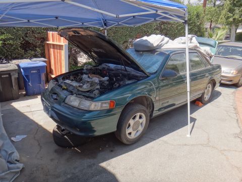1995 Ford Taurus - A green 1995 Ford Taurus sedan is parked under a blue canopy with its hood open, revealing the engine compartment.