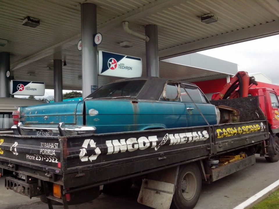 Blue sedan being transported on a flatbed truck at a gas station. Truck has 'INGOT METALS' written on the side