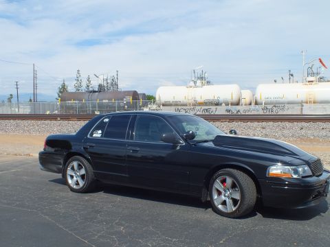 2003 Ford Crown Victoria - black full-size sedan on alloy wheels is parked along railroad tracks, with white tanker cars and graffiti behind under a blue sky.