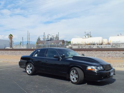 2003 Ford Crown Victoria - black sedan with tinted windows sits beside railroad tracks lined with tanker cars, under a blue sky with scattered clouds and palms.