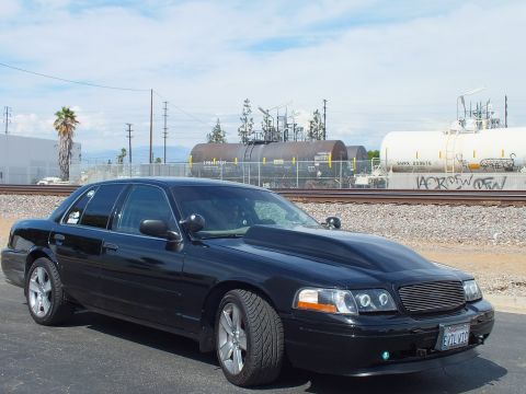 2003 Ford Crown Victoria - black sedan with a billet grille and halo headlights is parked near railroad tracks and tanker cars beneath a clear blue sky.