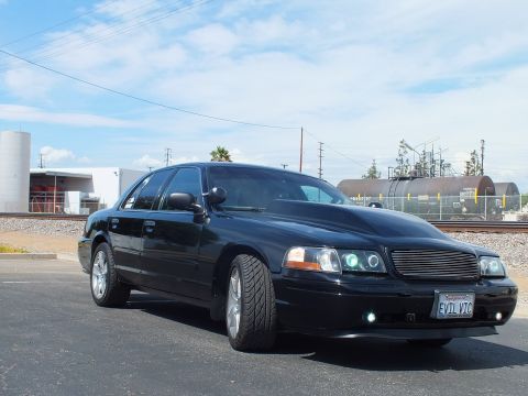 2003 Ford Crown Victoria - black sedan with a billet grille, halo headlights and an 'EVIL VIC' front plate is parked near railroad tanker cars under a blue sky.