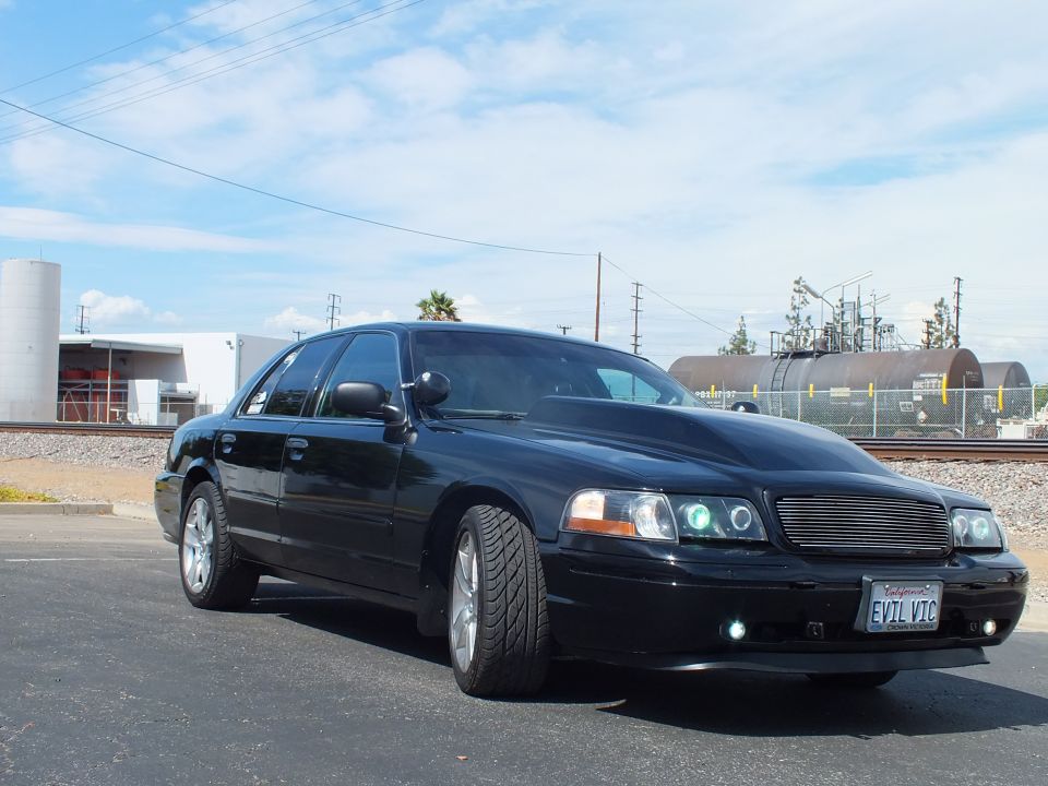 Black sedan with 'EVIL VIC' plate, billet grille and halo lights parked near rail tanker cars