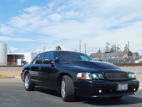 2003 Ford Crown Victoria - black full-size sedan with a billet grille, halo projector lights and an 'EVIL VIC' front plate sits by railroad tracks under blue sky.