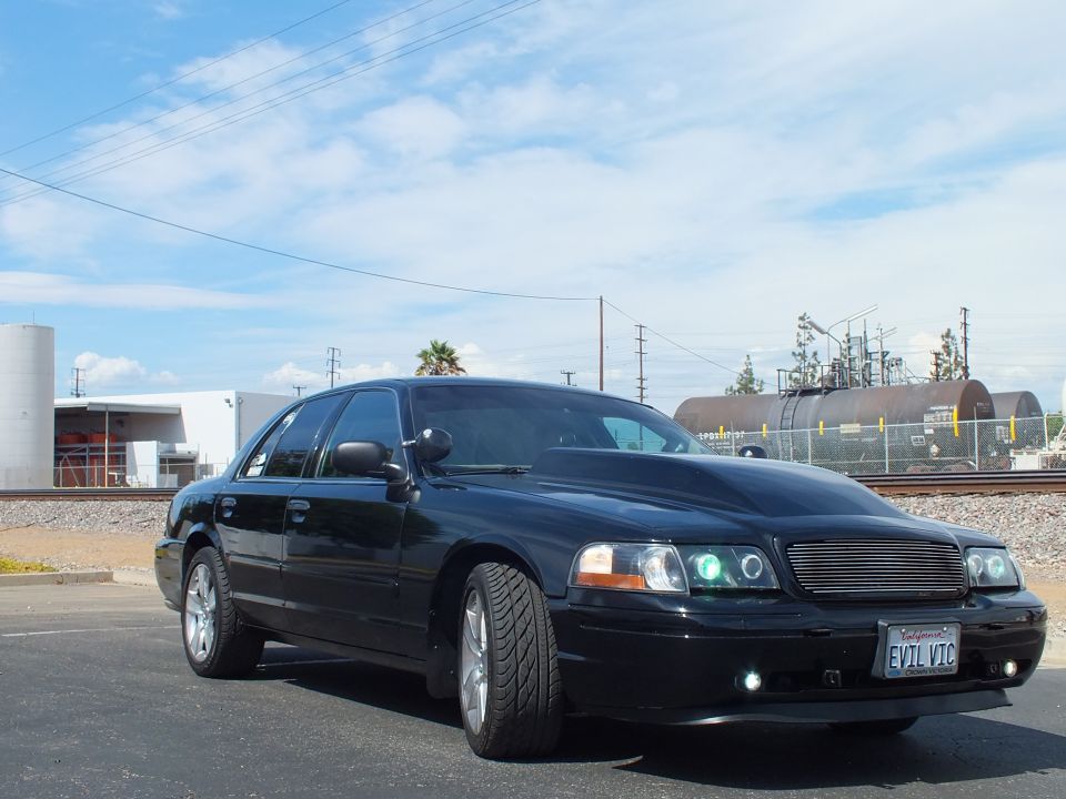 Front of a black sedan with billet grille, halo headlights and 'EVIL VIC' plate by rail tracks