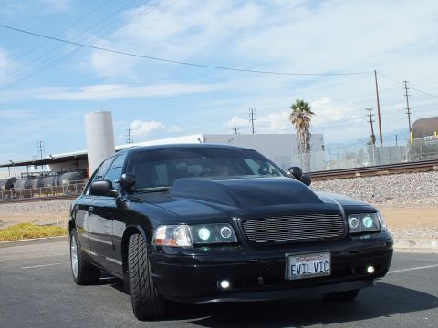 2003 Ford Crown Victoria - blacked-out full-size sedan with a billet grille, halo projector headlights and a California EVIL VIC plate, parked beside railroad tracks.