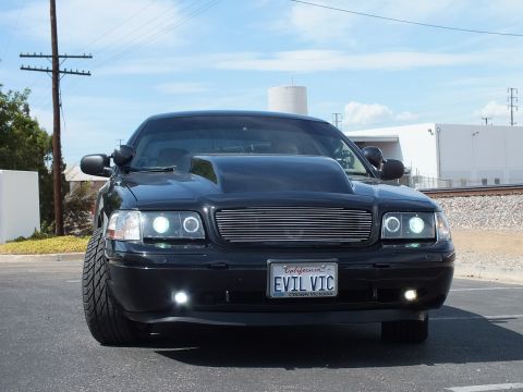 2003 Ford Crown Victoria - Head-on view of a lowered black sedan with a billet grille, glowing halo headlights and an EVIL VIC California plate in a parking lot.