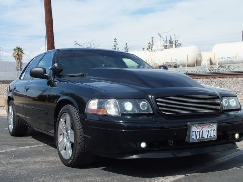 2003 Ford Crown Victoria - Front three-quarter view of a black sedan with chrome wheels and halo headlights parked near rail tank cars, wearing an EVIL VIC plate.