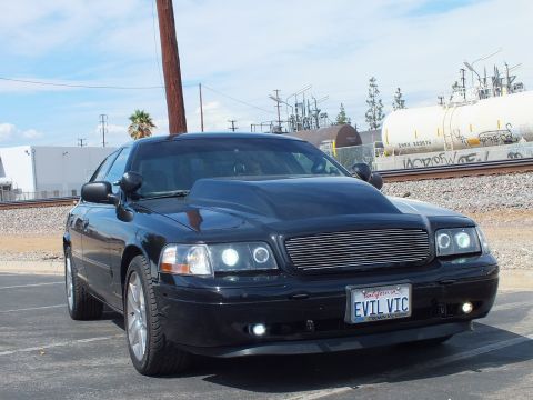 2003 Ford Crown Victoria - black sedan with a billet grille, halo lights and chrome wheels sits beside rail tank cars and power lines, plate reading EVIL VIC.