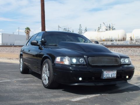 2003 Ford Crown Victoria - Front three-quarter shot of a black sedan with chrome wheels and a billet grille, EVIL VIC plate, parked by railroad tracks.